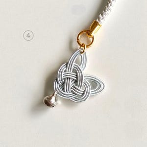 May include: A silver-colored cord charm featuring a decorative knot made of thin, metallic silver strands. The knot is attached to a gold-toned metal jump ring and a white braided cord. A small, round silver-colored bell hangs from the knot.