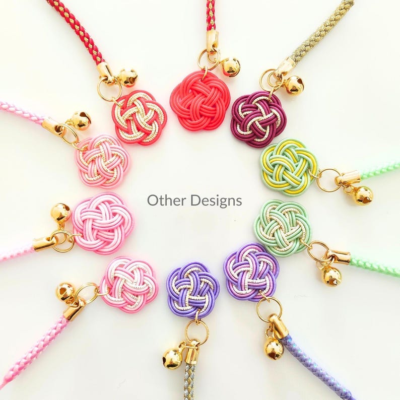 May include: A collection of ten decorative charms arranged in a circle, each featuring a knotted cord flower in various colors including red, pink, purple, green, and yellow, accented with gold-toned metal rings and small gold-toned bells.