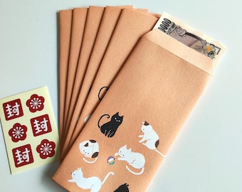 Puede incluir: Un juego de sobres de papel marrón claro con ilustraciones de gatos blancos y negros jugando con una pelota. Un sobre contiene un billete japonés de 1000 yenes. Se incluye una pequeña hoja de pegatinas con iconos florales rojos y el kanji 封.