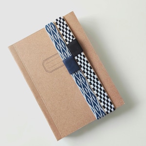 Könnte beinhalten: Ein beigefarbenes Notizbuch mit einem rechteckigen Etikettenumriss auf dem Einband, das von zwei elastischen Bändern gehalten wird. Ein Band hat einen blau-weißen Musterstoff mit dunkelblauer Mitte, das andere ein schwarz-weißes Karomuster mit schwarzer Mitte.
