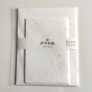 Puede incluir: Un juego de papelería con papel blanco de sutil textura fibrosa, sujeto por una banda de papel impresa con el texto japonés 伊予和紙 レターセット, que indica un contenido de seis hojas de papel para escribir y tres sobres.