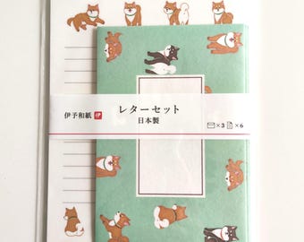 Puede incluir: Un juego de papelería en una funda de plástico transparente con sobres de color verde claro con un patrón de perros Shiba Inu y una banda de papel blanca con el texto "伊予和紙" y "レターセット 日本製". Incluye papel de carta con líneas.