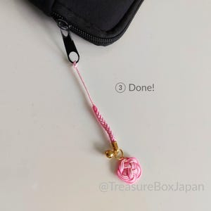 May include: A pink cord charm attached to a black zipper pull. The charm features a braided pink cord, a small gold-colored bell, and a pink knotted flower-shaped ornament. The text 3 Done! and @TreasureBoxJapan are visible in the image.