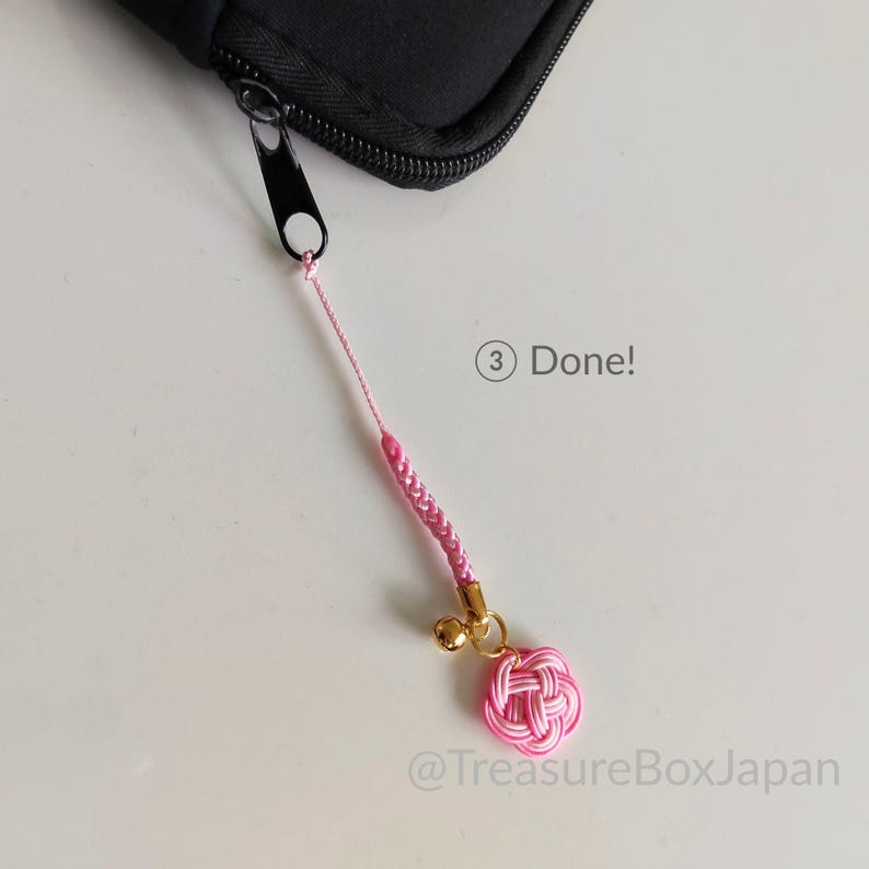 May include: A pink cord charm attached to a black zipper pull. The charm features a braided pink cord, a small gold-colored bell, and a pink knotted flower-shaped ornament. The text 3 Done! and @TreasureBoxJapan are visible in the image.
