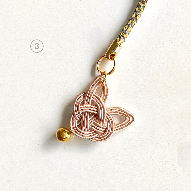 May include: A pale pink mizuhiki knot charm shaped like a rabbit head with a small gold bell attached to the bottom. The charm is connected by a gold jump ring to a braided gold and silver cord. The number 3 is visible in a circle on the left.