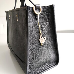 May include: A black pebbled leather tote bag featuring a gold-toned metal ring on the side. Attached to the ring is a decorative charm made of light-colored cord tied in a knot, accented with a small gold bead and a metallic cylindrical component.