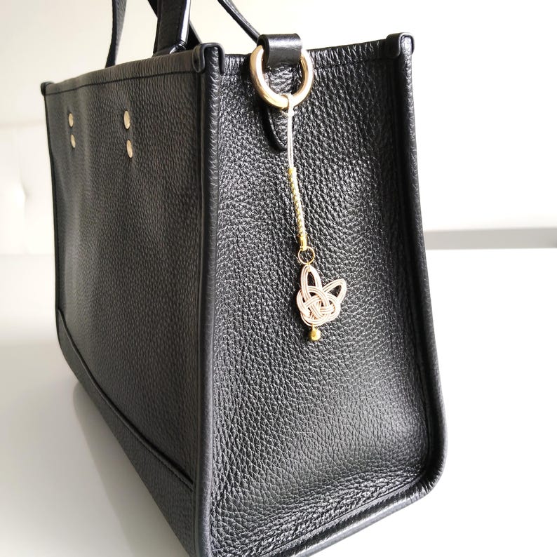 May include: A black pebbled leather tote bag featuring a gold-toned metal ring on the side. Attached to the ring is a decorative charm made of light-colored cord tied in a knot, accented with a small gold bead and a metallic cylindrical component.