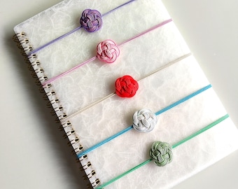 May include: A spiral-bound notebook with a textured white cover featuring five elastic bands, each adorned with a decorative knot in purple, pink, red, white, and green. The bands are arranged horizontally across the notebook cover.