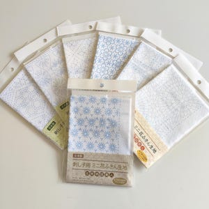 May include: A collection of six white fabric squares for sashiko embroidery, each packaged in a clear plastic sleeve. The fabric features various blue geometric and floral patterns printed on the surface. Each package includes Japanese text labels.