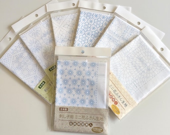 Puede incluir: Una colección de seis cuadrados de tela blanca para bordado sashiko, cada uno empaquetado en una funda de plástico transparente. La tela presenta varios patrones geométricos y florales azules impresos. Cada paquete incluye etiquetas con texto en japonés.