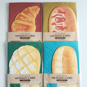 May include: Four rectangular message cards in clear plastic sleeves. Each card features a die-cut bread illustration: a golden croissant on red, a hot dog on gold, a melon pan on green, and a cream-filled bun on teal. Each has the text MESSAGE CARD.