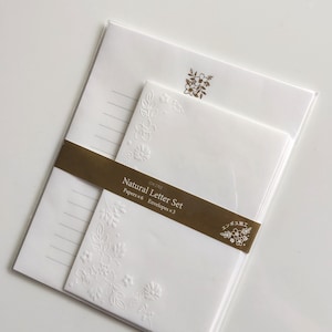 Może przedstawiać: Biały zestaw papeterii zawierający sześć liniowanych kartek i trzy koperty spięte brązową papierową opaską z napisem Natural Letter Set. Papier posiada subtelny tłoczony wzór kwiatowy w rogu oraz małą złotą ikonę kwiatka na górze.