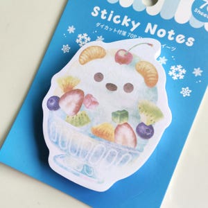 Op de afbeelding: Een gestanst memoblok met een aquarelillustratie van een wit, beervormig dessert in een blauwe kom, gegarneerd met diverse vruchten zoals aardbeien, bosbessen en sinaasappelpartjes op een blauwe achtergrondkaart met sneeuwvlokpictogrammen.
