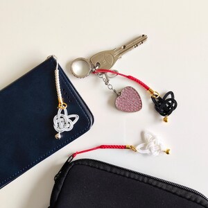 May include: Three decorative charms featuring intricate knotted cord designs in white and black, attached to red or white braided cords with small gold-toned beads. One charm is attached to a silver key ring with a pink heart-shaped pendant.