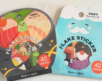 Puede incluir: Dos paquetes de pegatinas tipo flake, cada uno con 40 piezas. El paquete izquierdo presenta diseños de sushi sobre un fondo gris oscuro. El paquete derecho presenta diseños de personajes sobre un fondo azul claro. Ambos paquetes están marcados como Made in Japan.