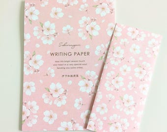 Puede incluir: Una hoja de papel de carta rectangular y un sobre a juego, ambos con un fondo rosa suave y un patrón repetitivo de flores de cerezo blancas. El papel incluye el texto "Sakuramaguri WRITING PAPER" y caracteres japoneses.