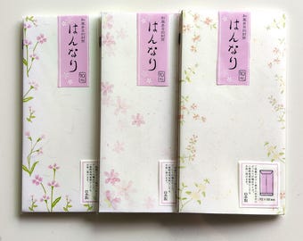 Puede incluir: Tres sobres de papel rectangulares con delicados motivos florales en tonos rosa y verde. Cada sobre tiene una etiqueta vertical morada con texto en japonés y el número 10, y una etiqueta blanca pequeña que indica un tamaño de 192 x 100 mm.