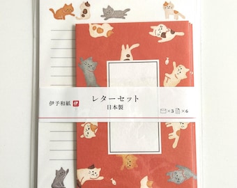 Puede incluir: Un juego de papelería en embalaje transparente que incluye papel blanco rayado con ilustraciones de gatos pequeños y una pila de sobres rojos decorados con varios patrones de gatos. Una banda de papel blanca en el centro muestra el texto "伊予和紙" y "レターセット 日本製" con iconos que indican tres sobres y seis hojas de papel.