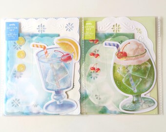 May include: Two amifa letter sets in clear plastic packaging. The left set features a blue drink with ice, a cherry, and a lemon slice. The right set features a green drink with ice, a cherry, and a scoop of ice cream. Both include decorative paper.