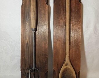 May include: Two wooden wall hangings featuring kitchen utensils. One has a metal fork with a wooden handle, the other a wooden spoon. Both are mounted on rectangular, stained wooden plaques, showcasing a rustic, farmhouse aesthetic.