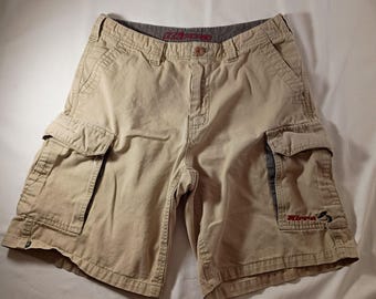 May include: Beige cargo shorts featuring a button and zipper fly closure, two side cargo pockets with flaps, and two front slash pockets. The interior waistband displays the Kirra brand name in red text, which is also embroidered on the right pocket.