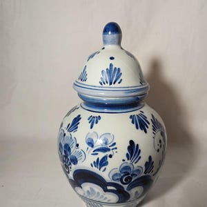 May include: A white and blue ceramic jar with a lid. The jar is decorated with blue floral patterns and a blue finial on the lid. The jar is a round shape and has a blue band around the middle.