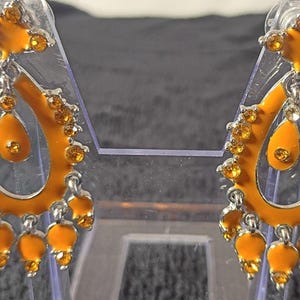 以下が含まれることがあります： オレンジとシルバーカラーのドロップイヤリングのペア。イヤリングは、オレンジ色の宝石のクラスターと単一のティアドロップ宝石を備えたティアドロップデザインです。イヤリングは透明なディスプレイスタンドにあります。