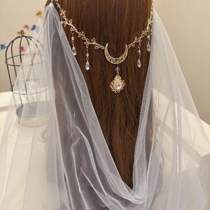 May include: A gold-colored headpiece with a crescent moon and teardrop-shaped crystal pendants. The headpiece is worn with a white veil. The hair is brown.