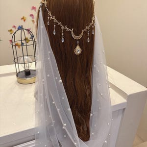 May include: A gold-colored headpiece with a crescent moon and dangling crystals is worn over brown hair. A white veil with scattered pearls drapes down. The headpiece is part of a bridal or special occasion ensemble.