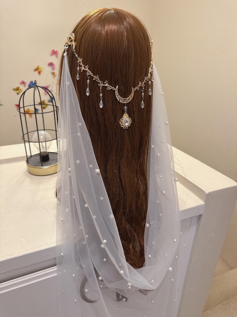 May include: A gold-colored headpiece with a crescent moon and dangling crystals is worn over brown hair. A white veil with scattered pearls drapes down. The headpiece is part of a bridal or special occasion ensemble.