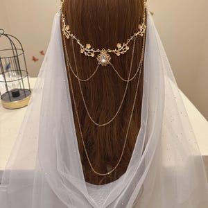 May include: A gold-colored bridal headpiece with floral and chain details, featuring a central teardrop-shaped gem. A sheer white veil with subtle sparkle drapes down. The headpiece is worn on brown hair.