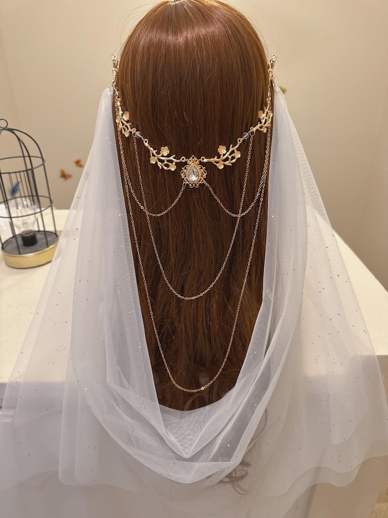 May include: A gold-colored bridal headpiece with floral and chain details, featuring a central teardrop-shaped gem. A sheer white veil with subtle sparkle drapes down. The headpiece is worn on brown hair.