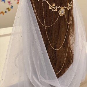 May include: A gold-colored headpiece with floral and chain details, adorned with a central gemstone. The headpiece is worn with a white, sheer veil that drapes over the hair. The background is blurred, with hints of colorful butterfly decorations.