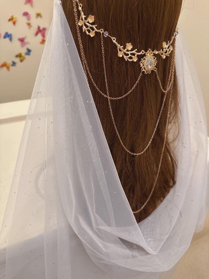 May include: A gold-colored headpiece with floral and chain details, adorned with a central gemstone. The headpiece is worn with a white, sheer veil that drapes over the hair. The background is blurred, with hints of colorful butterfly decorations.