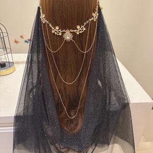May include: A gold-colored headpiece with a floral design and a central jewel, paired with a black, sparkly veil. The headpiece features delicate chains that drape down. The veil is a sheer fabric with a subtle shimmer.