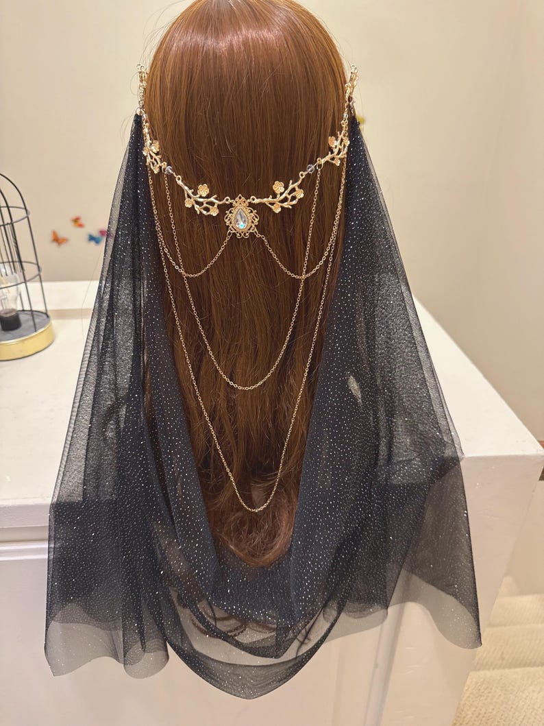 May include: A gold-colored headpiece with a floral design and a central jewel, paired with a black, sparkly veil. The headpiece features delicate chains that drape down. The veil is a sheer fabric with a subtle shimmer.