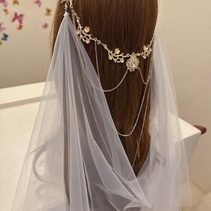 May include: A gold-colored bridal headpiece with a floral design and dangling chains. The headpiece is adorned with a large, clear gemstone and is paired with a sheer white veil. The headpiece is designed to be worn on the head.