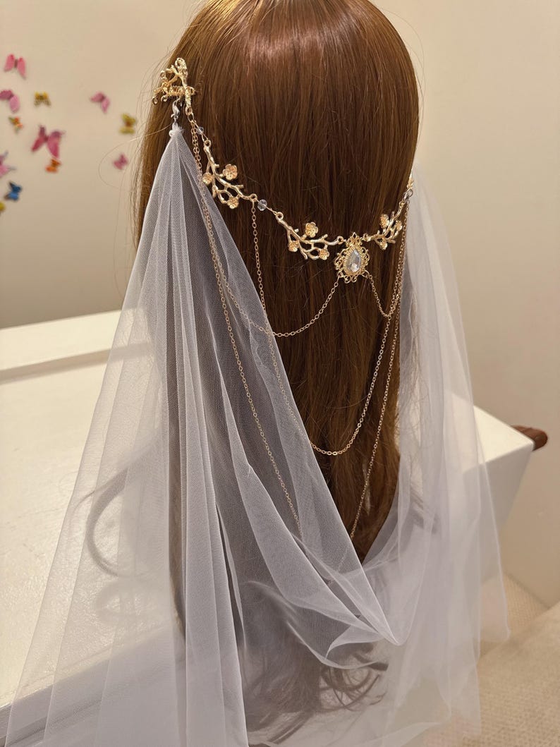 May include: A gold-colored bridal headpiece with a floral design and dangling chains. The headpiece is adorned with a large, clear gemstone and is paired with a sheer white veil. The headpiece is designed to be worn on the head.