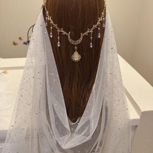May include: A gold-toned celestial headpiece with a crescent moon and dangling crystals. The headpiece is worn with a white, sheer veil sprinkled with iridescent glitter. The design is elegant and suitable for weddings or special occasions.