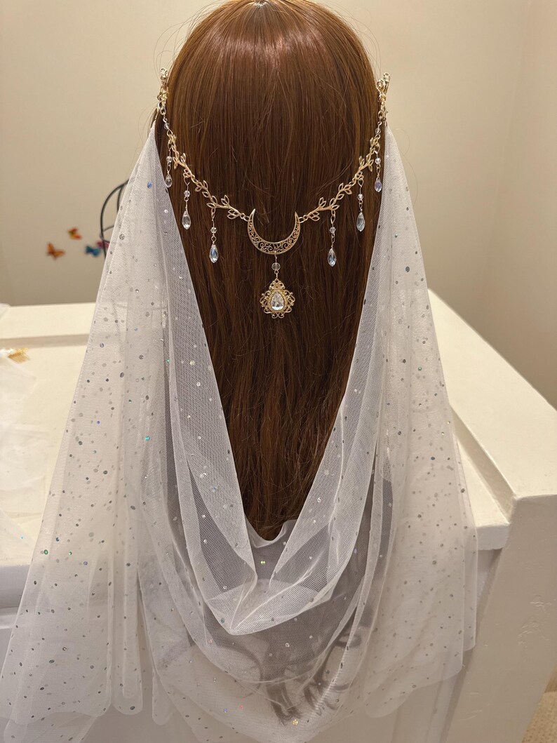 May include: A gold-toned celestial headpiece with a crescent moon and dangling crystals. The headpiece is worn with a white, sheer veil sprinkled with iridescent glitter. The design is elegant and suitable for weddings or special occasions.
