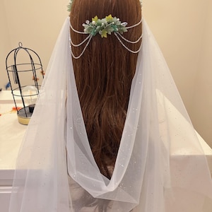 Op de afbeelding: Bruidssieraden met een bloemenkrans met groene en gele accenten, verbonden met een ketting die over het haar valt. Een doorschijnende witte sluier met glinsterende details is over het hoofd gedrapeerd.