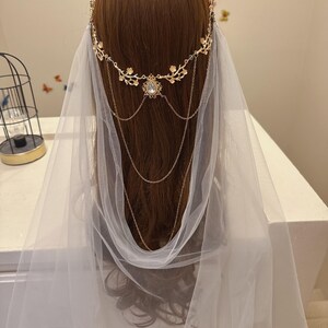 May include: A gold-colored bridal headpiece with floral accents and dangling chains, paired with a sheer white veil. The headpiece features a central jewel and delicate detailing, creating an elegant and romantic look. The veil drapes softly.