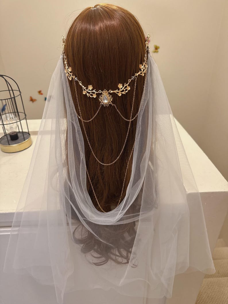 May include: A gold-colored bridal headpiece with floral accents and dangling chains, paired with a sheer white veil. The headpiece features a central jewel and delicate detailing, creating an elegant and romantic look. The veil drapes softly.