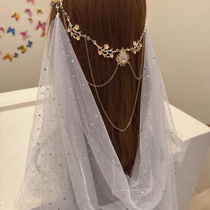 May include: A gold-colored headpiece with floral and chain details, paired with a sheer white veil. The headpiece features a central jewel and delicate chain accents, creating an elegant, ethereal look. The veil is adorned with subtle, shimmering embellishments.
