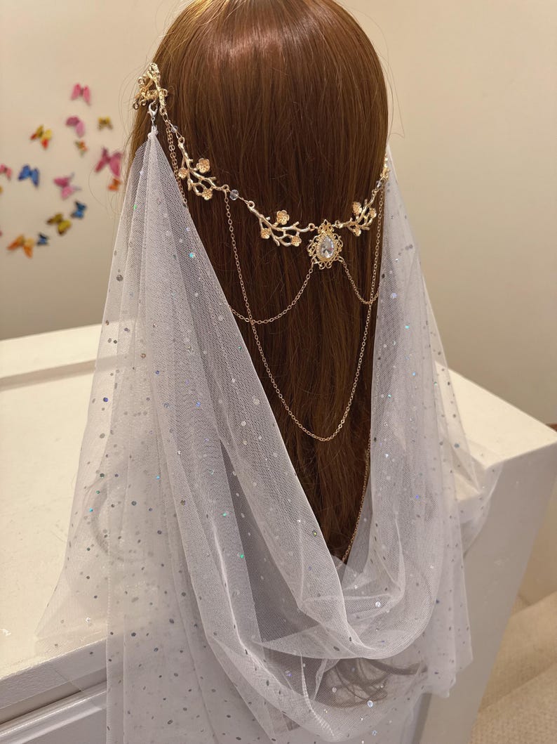 May include: A gold-colored headpiece with floral and chain details, paired with a sheer white veil. The headpiece features a central jewel and delicate chain accents, creating an elegant, ethereal look. The veil is adorned with subtle, shimmering embellishments.