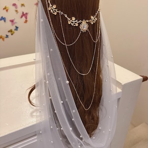 May include: A bridal headpiece with a gold floral crown and cascading chains. A sheer white veil is adorned with scattered pearls. The headpiece is set against brown hair.
