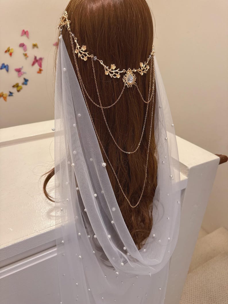 May include: A bridal headpiece with a gold floral crown and cascading chains. A sheer white veil is adorned with scattered pearls. The headpiece is set against brown hair.