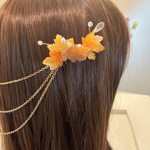 Op de afbeelding: Een haaraccessoire met amberkleurige bladvormige versieringen, heldere kristallen accenten en kleine witte kralen. Goudkleurige kettingen hangen van de bladeren af. Het haarstuk is ontworpen om in het haar te worden gedragen.