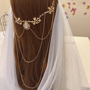 May include: A gold-colored bridal headpiece with floral and chain details. The headpiece features a central teardrop-shaped gem and cascading chains. A sheer white veil is draped behind the headpiece, creating a soft, ethereal look.