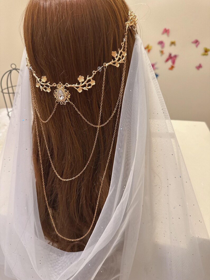 May include: A gold-colored bridal headpiece with floral and chain details. The headpiece features a central teardrop-shaped gem and cascading chains. A sheer white veil is draped behind the headpiece, creating a soft, ethereal look.
